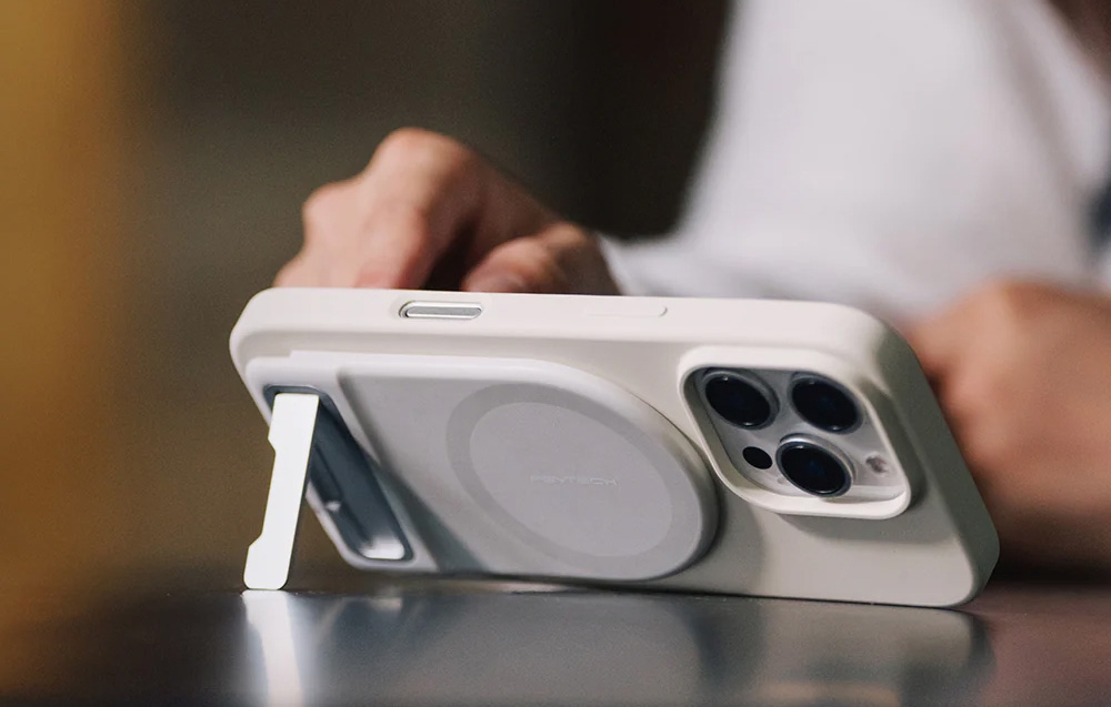 iPhone in a white case with a MagSafe power bank and built-in stand set vertically on a table, hands in the background