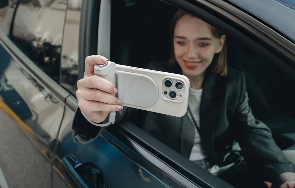 Woman in a car holding an iPhone in a white case with a side grip and power bank, taking a selfie