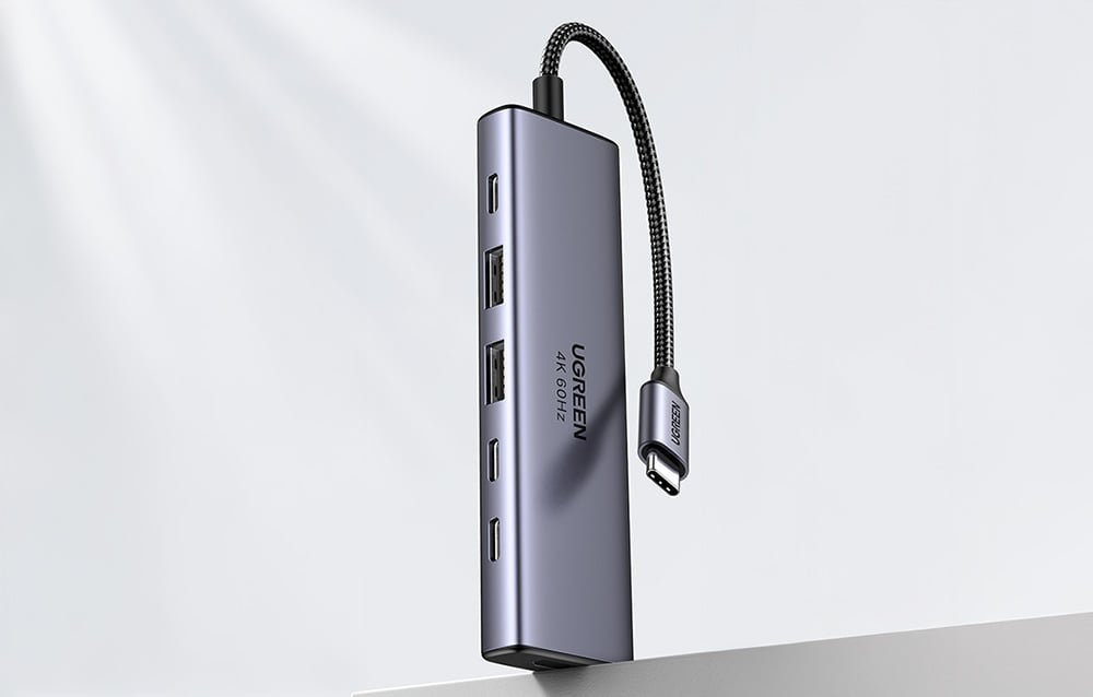Hub UGREEN pokazany pionowo nad krawędzią stołu z widocznym przewodem USB-C i zestawem portów.
