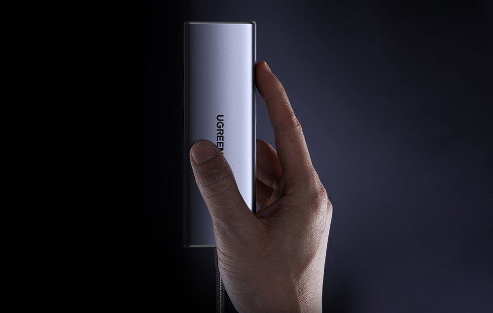 Osoba trzymająca hub UGREEN pionowo między palcami na ciemnym tle.