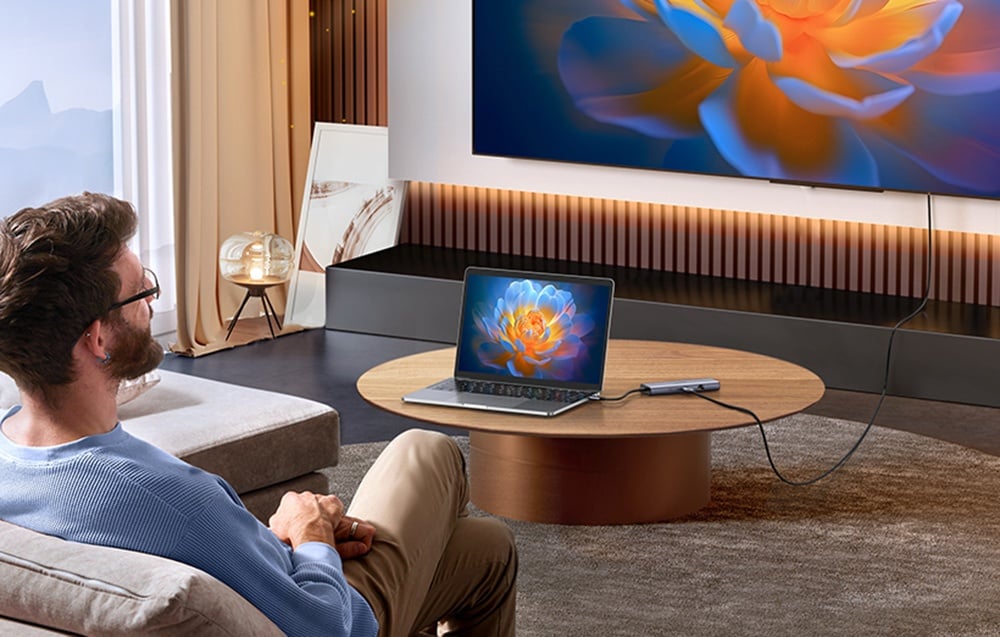 Mężczyzna siedzący na kanapie w salonie ogląda treści z laptopa podłączonego przez hub UGREEN do dużego telewizora.