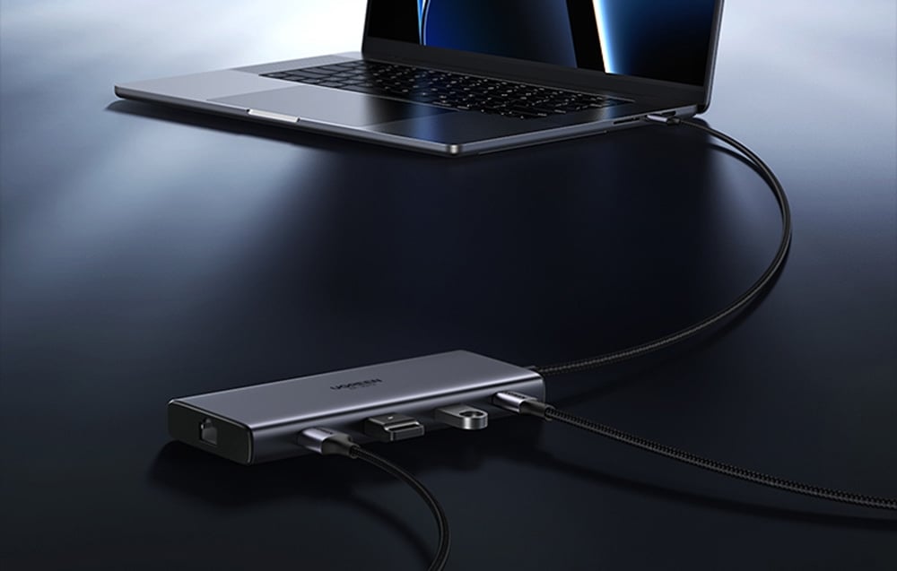Hub UGREEN podłączony do laptopa z wieloma kablami i akcesoriami, leżący na ciemnej powierzchni.
