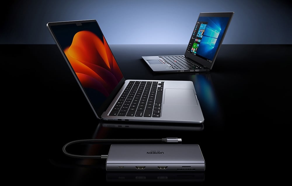 Hub UGREEN z portami HDMI i czytnikiem kart podłączony do laptopa, w tle drugi laptop z systemem Windows.