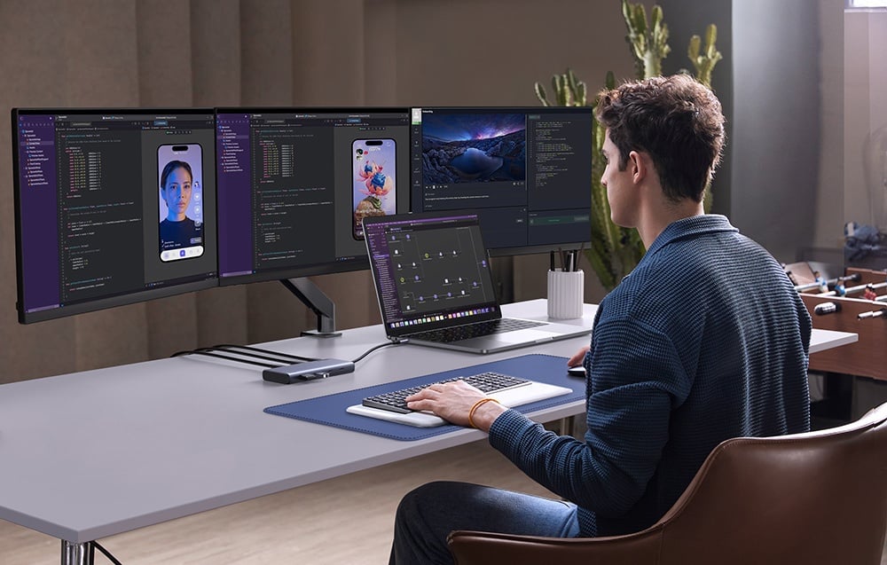 Mężczyzna pracujący przy laptopie i trzech monitorach z aplikacjami deweloperskimi, z użyciem huba UGREEN.