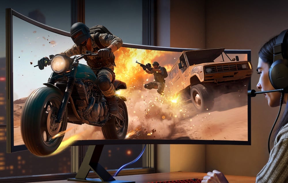 Monitor, z którego wyjeżdża motocyklista i duży pojazd; przy biurku kobieta w słuchawkach.