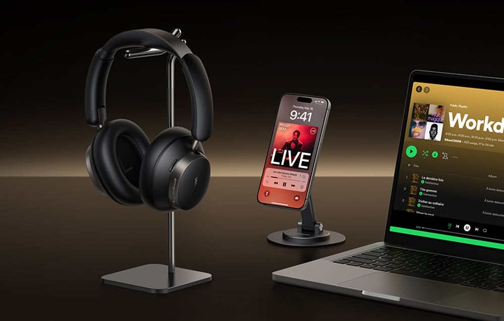 czarne słuchawki nauszne na stojaku obok telefonu w uchwycie i laptopa z włączonym Spotify