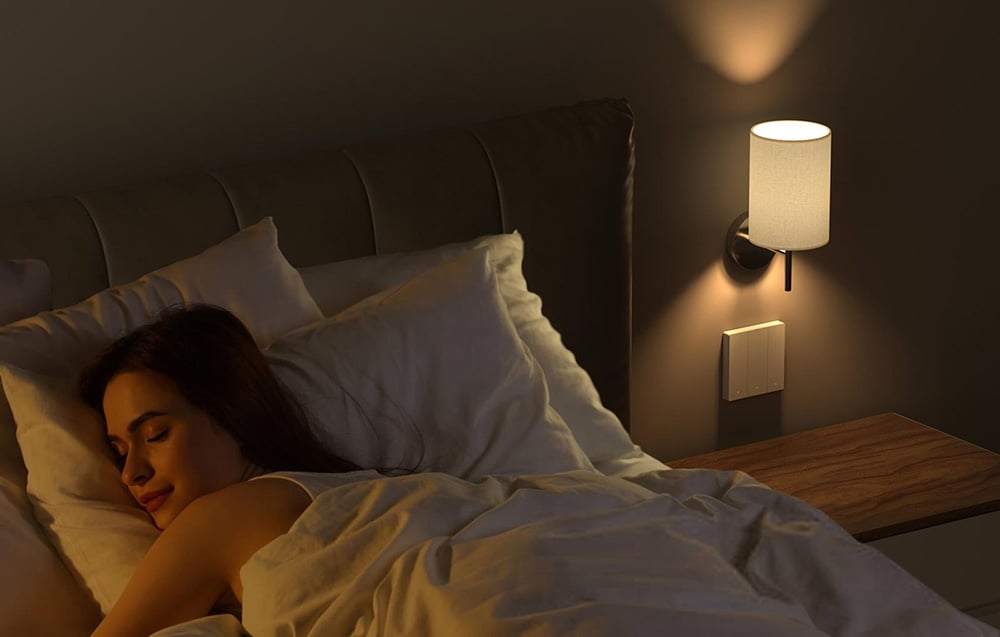 Kobieta śpiąca w łóżku z białą pościelą przy ścianie z inteligentnym przełącznikiem i lampką nocną