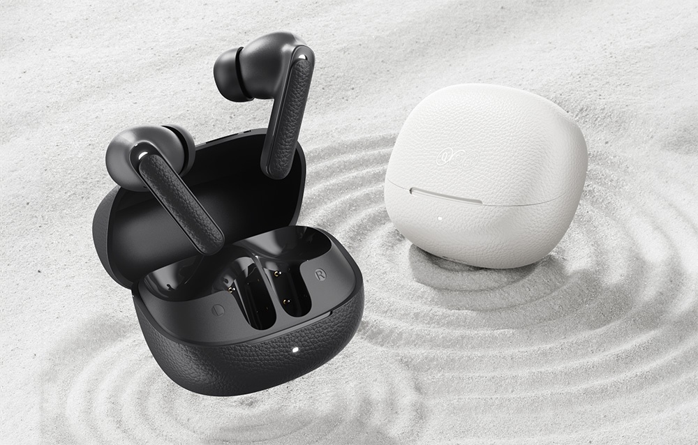 Czarne bezprzewodowe słuchawki douszne w etui ładującym obok zamkniętego białego etui, ustawione na jasnym piasku.