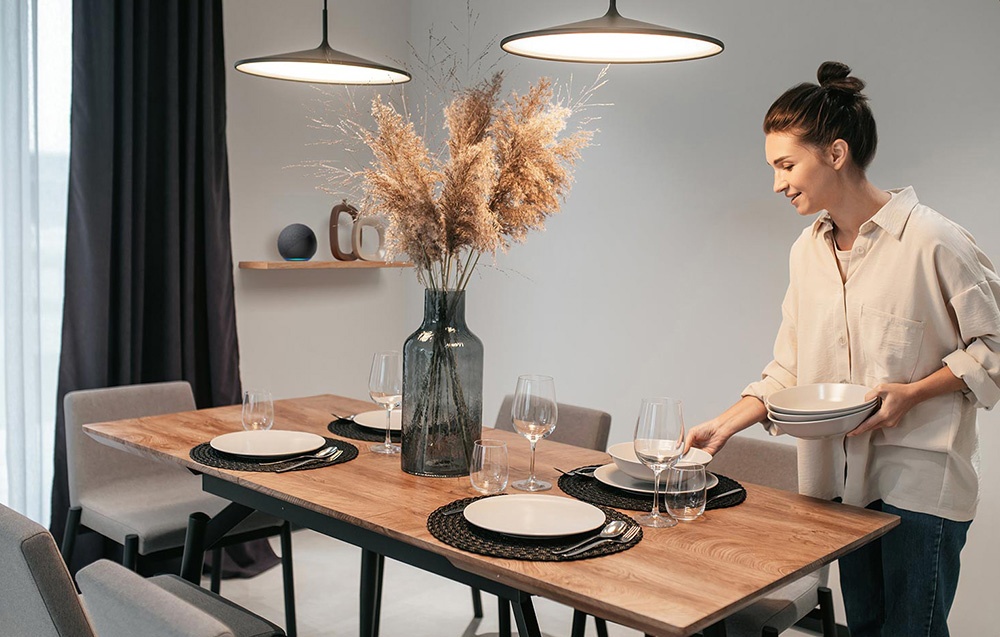 Kobieta w beżowej koszuli nakrywa stół z drewnianym blatem, szarymi talerzami i trawą pampasową w wazonie