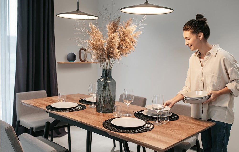 Kobieta w beżowej koszuli nakrywa stół z drewnianym blatem, szarymi talerzami i trawą pampasową w wazonie