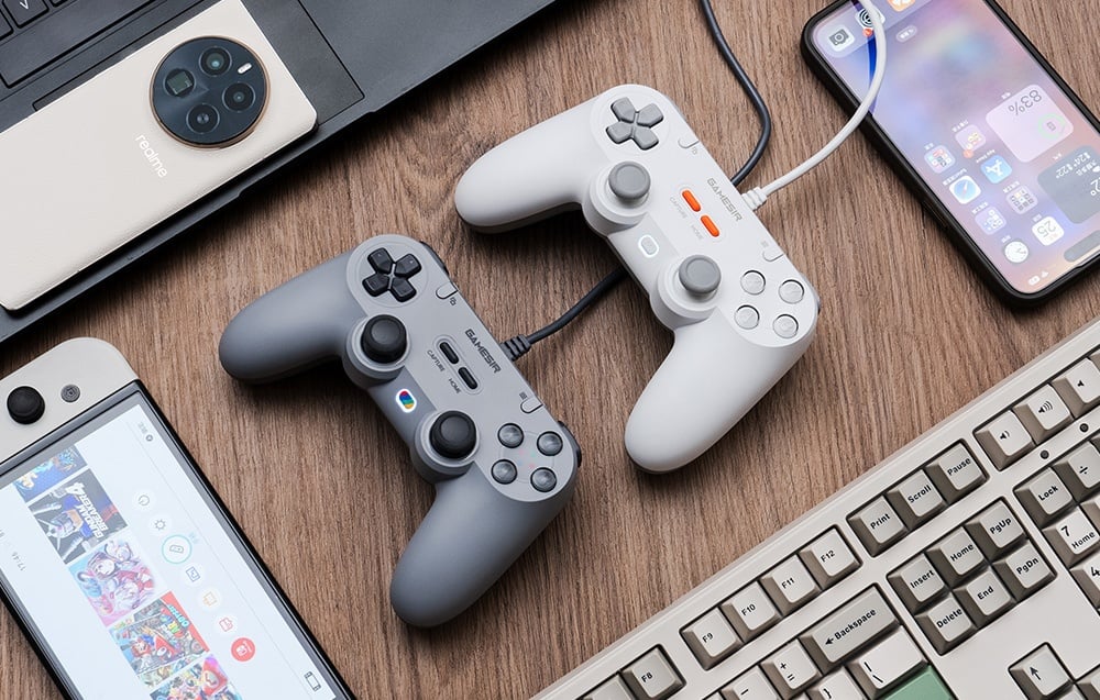 Dwa kontrolery GameSir, biały i szary, leżące na drewnianym biurku wśród różnych urządzeń elektronicznych.