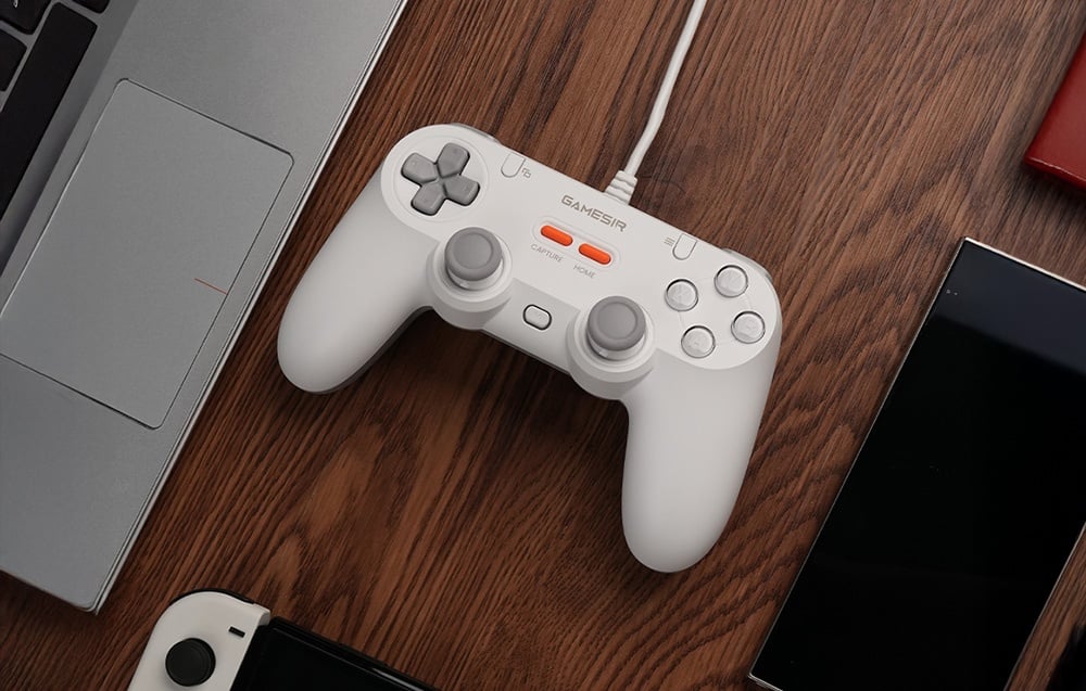 Biały kontroler GameSir podłączony kablem, ułożony na drewnianym blacie obok laptopa i smartfonów.