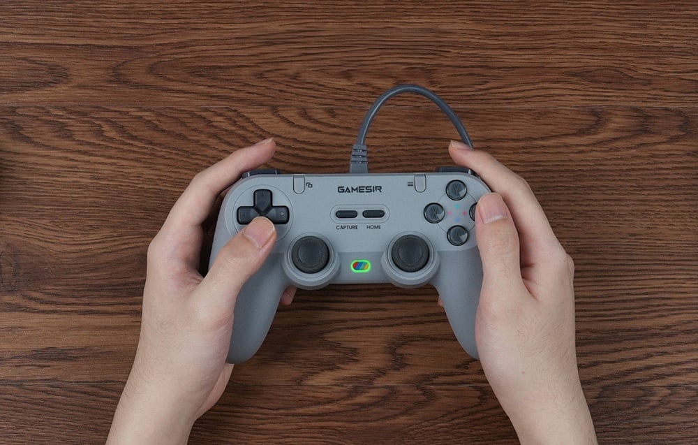 Osoba trzymająca szary kontroler GameSir obiema rękami nad drewnianym stołem.