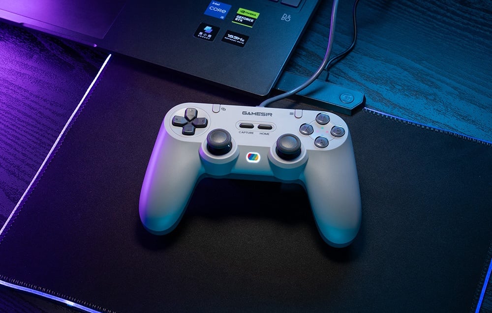 Szary kontroler GameSir leżący na podkładce przed laptopem, oświetlony niebiesko-fioletowym światłem.