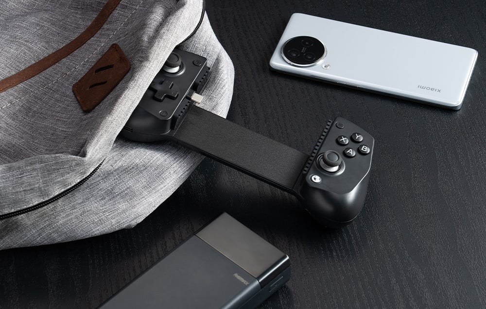 Czarny kontroler GameSir częściowo wystający z plecaka, obok smartfona i powerbanka na czarnym stole.