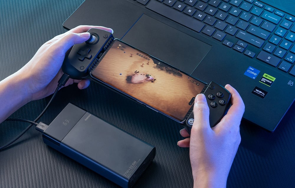 Osoba grająca na smartfonie zamontowanym w czarnym kontrolerze GameSir, z podłączonym powerbankiem obok laptopa.