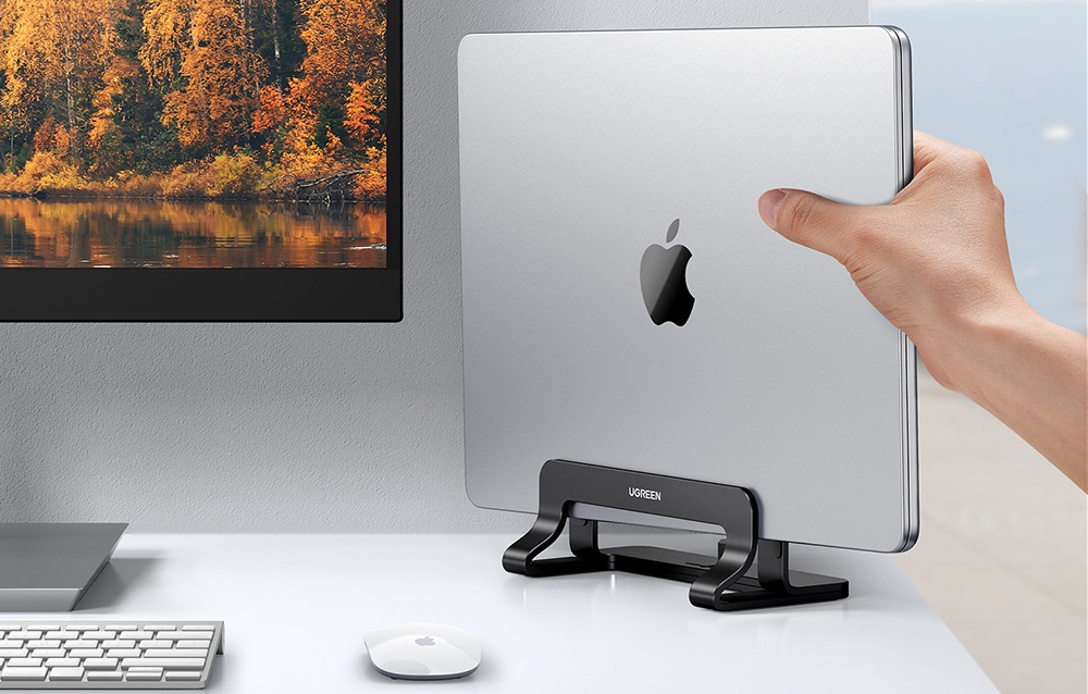 A hand placing a MacBook in a black UGREEN stand at a desk with a monitor and keyboard