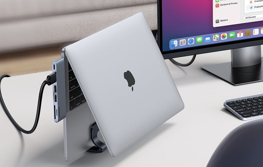 A MacBook in a stand connected by cable to a docking station on a white desk with a monitor and keyboard