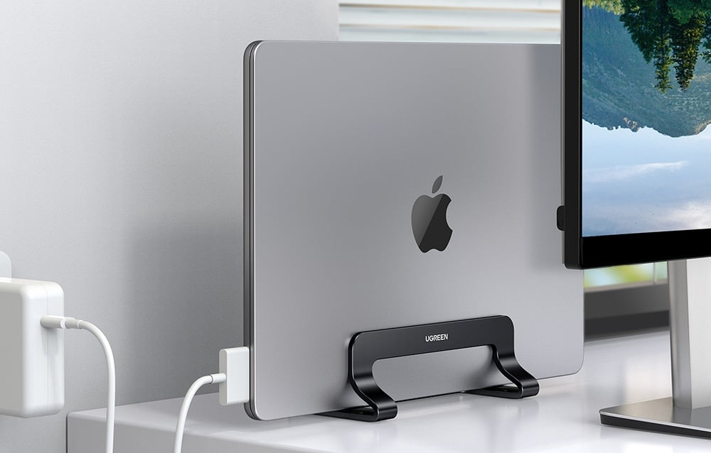 A MacBook in a black UGREEN stand next to a monitor and charger on a modern white desk