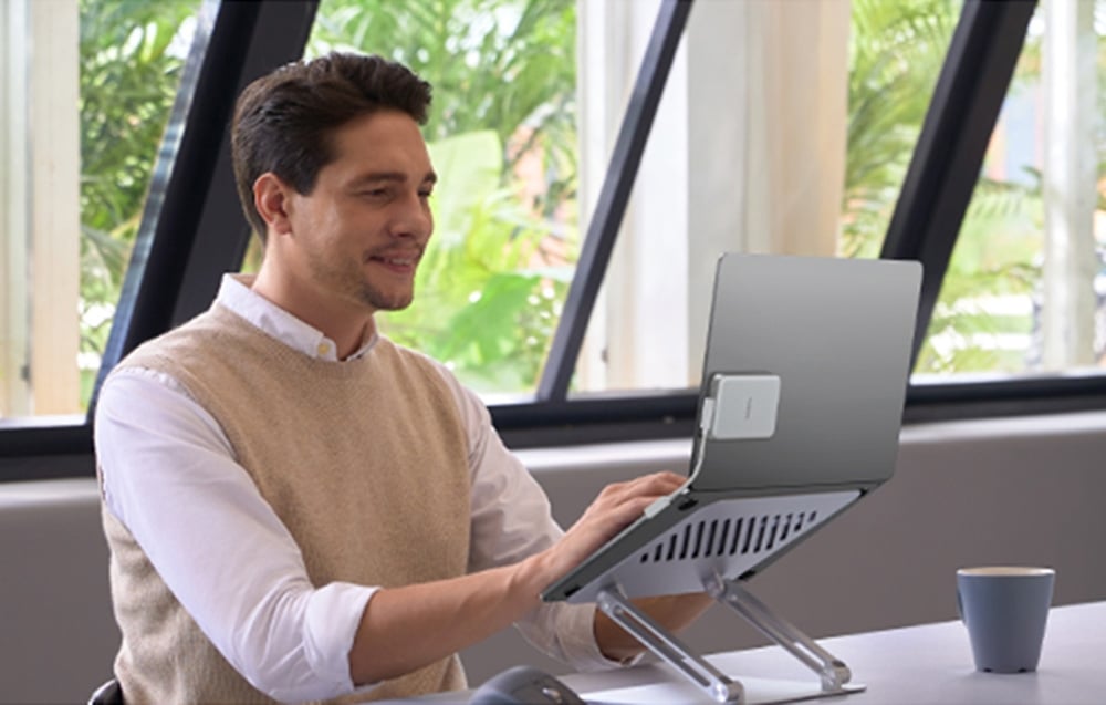 A man in a beige vest working on a laptop with a stand in an office by the window