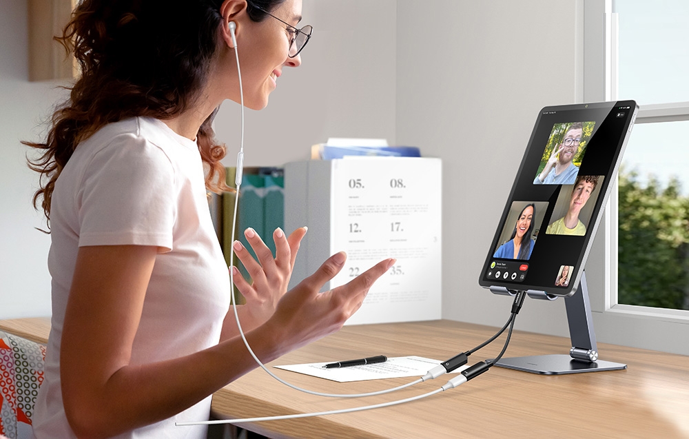 A woman in glasses conducts a video conference on a tablet connected to cables, gesturing at a desk