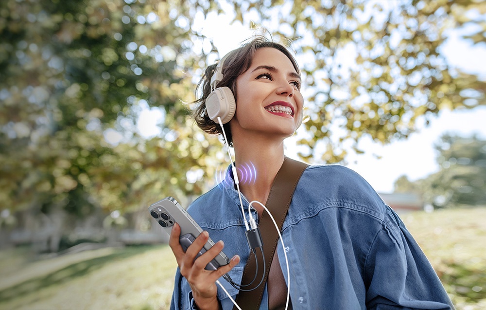 A smiling woman in a blue denim jacket listens to music through headphones while holding a phone in a park