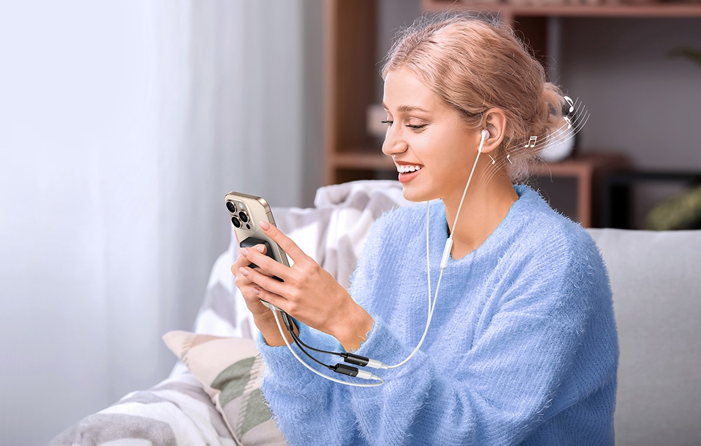 A blonde woman in a blue sweater listens to music through headphones, looking at her phone while sitting on a couch