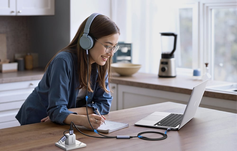 A woman wearing glasses and headphones studies at a laptop in the kitchen, taking notes in a notebook