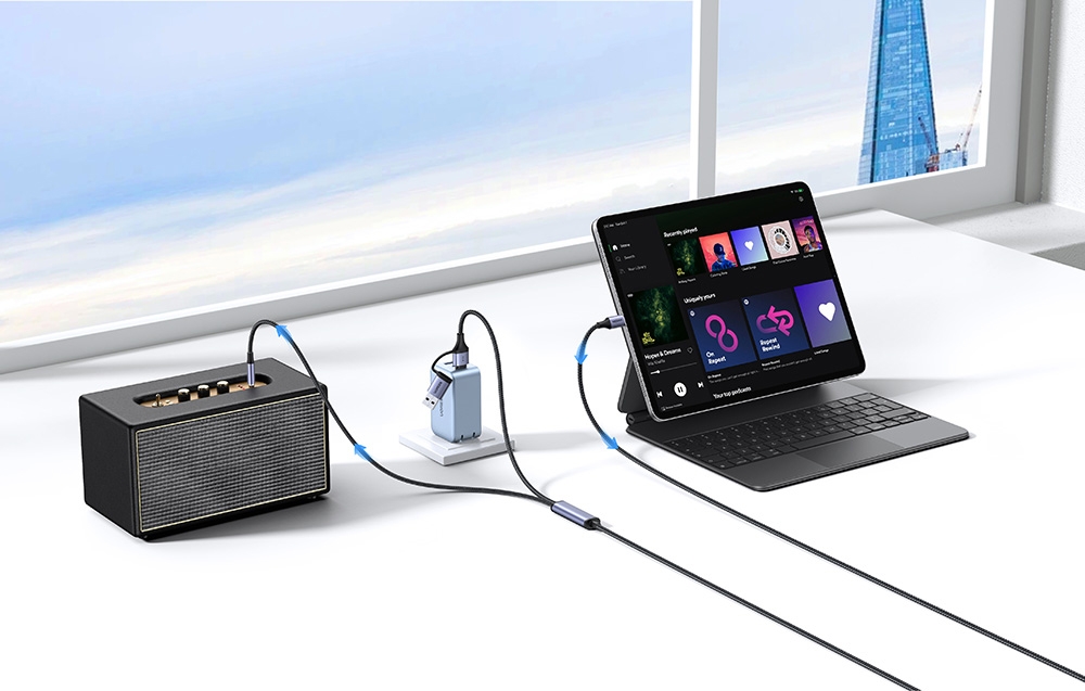 A laptop with a music application connected by cables to a speaker and charger on a white desk by the window