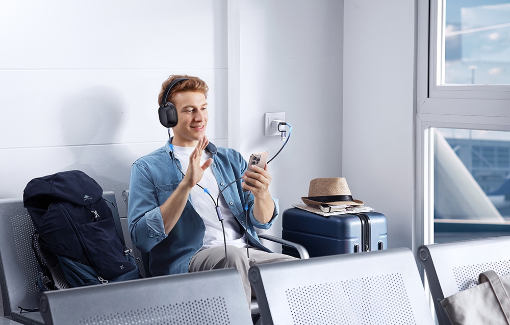 A smiling man in headphones charges his phone in an airport waiting area with luggage and a suitcase