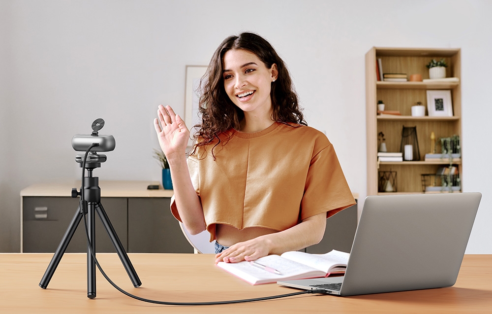 A smiling woman in an orange blouse waves during a video conference with the camera on a tripod