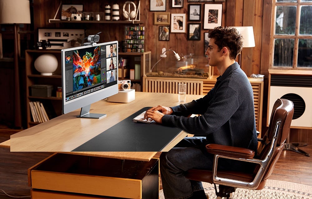 A man in a gray sweater works at an iMac computer with a camera in a cozy wooden office