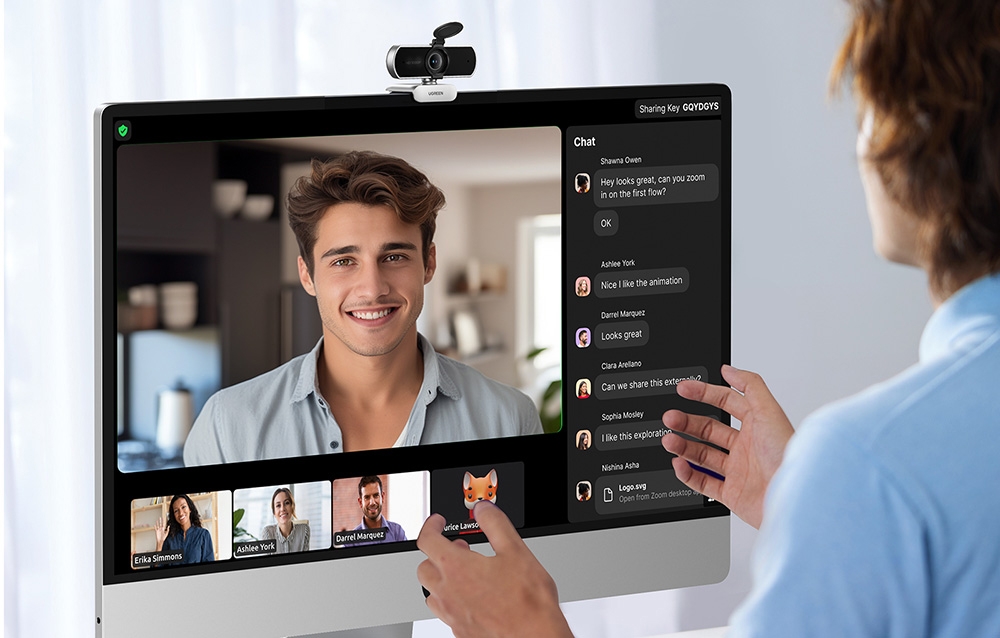 Person in a blue shirt gesturing during a video conference on a monitor with a camera and chat participants on the right side