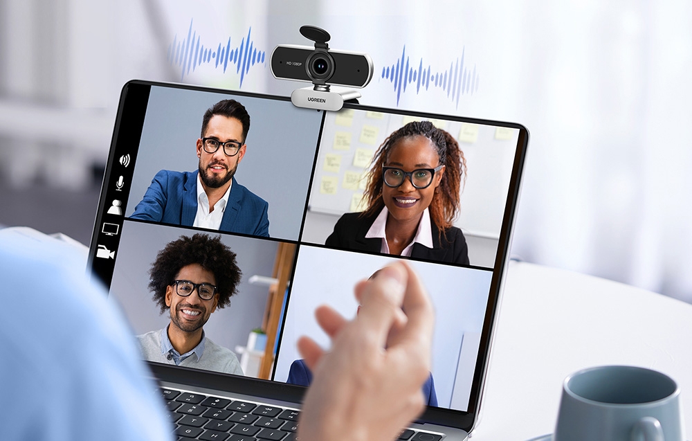 Laptop with a video conference of four participants, camera on top, and a hand gesturing towards the screen