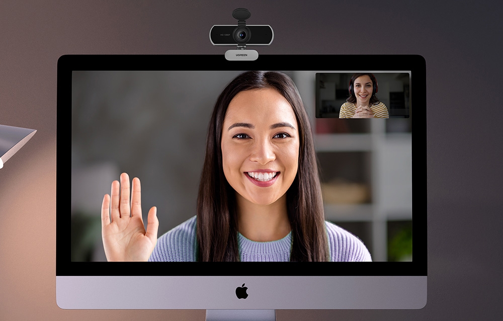 Smiling woman in a gray sweater waving during a video call on iMac with UGREEN camera