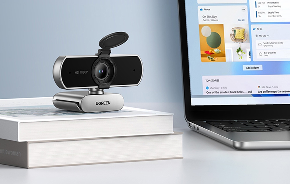 UGREEN webcam with a privacy cover on white books next to a laptop with an application