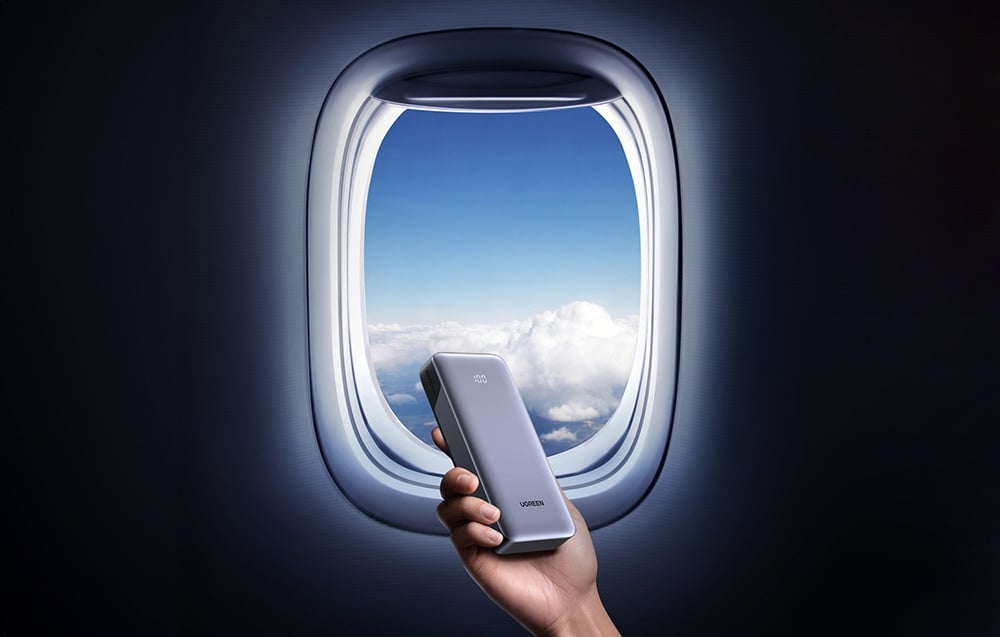 Ręka trzymająca powerbank UGREEN przy oknie samolotu z widokiem na niebo