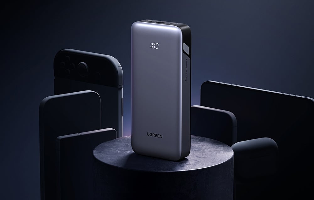 Powerbank UGREEN ustawiony pionowo na cylindrycznym postumencie w otoczeniu ciemnych smartfonów