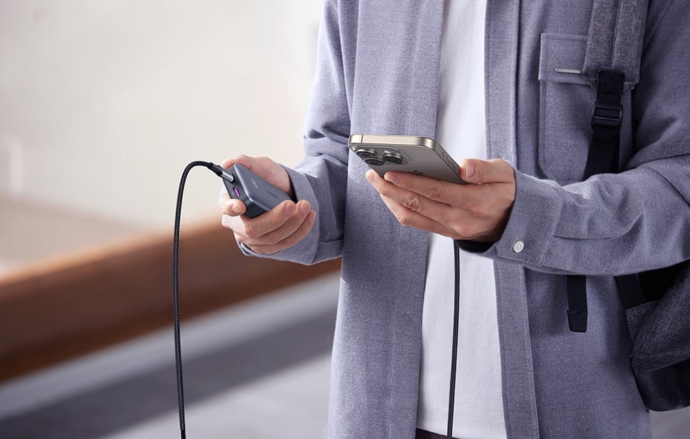 A man in a gray jacket holding a power bank and smartphone connected by a cable