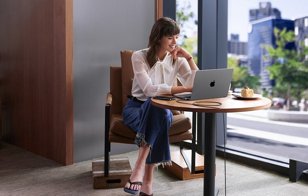 A woman in a white shirt working on a MacBook at a table in a café with a city view