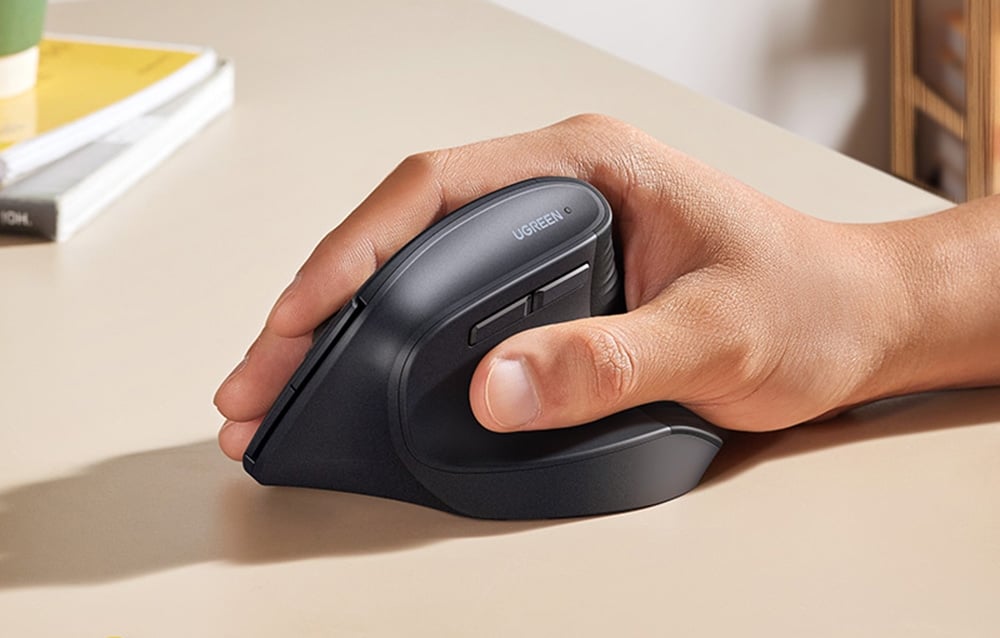 Zbliżenie dłoni trzymającej pionową myszkę UGREEN na blacie biurka
