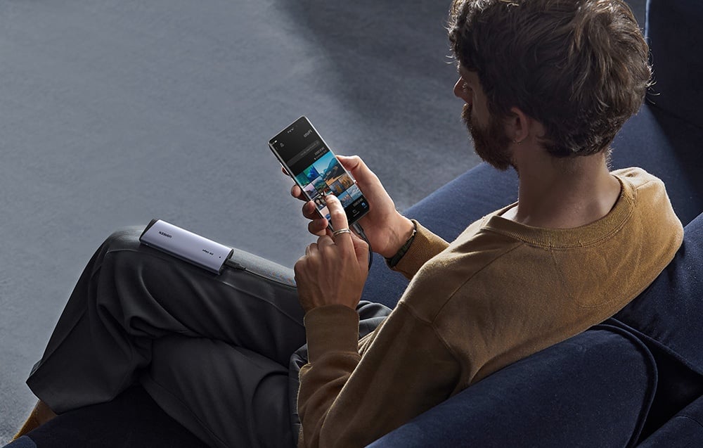 A man sitting on a couch browsing photos on a smartphone connected to the UGREEN drive