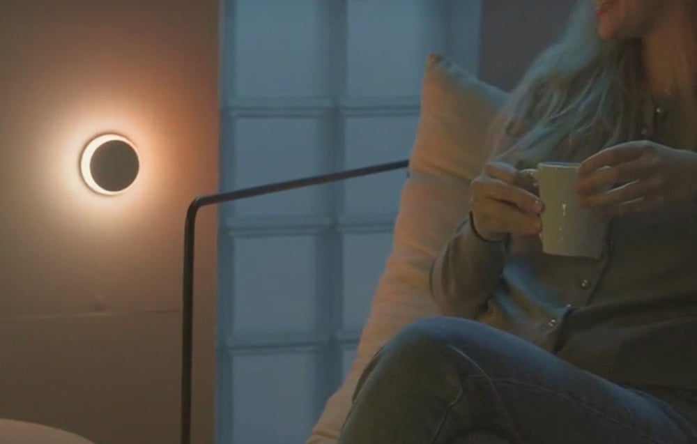 Lampka nocna Yeelight Vega świecąca ciepłym światłem na ścianie obok siedzącej osoby trzymającej kubek.