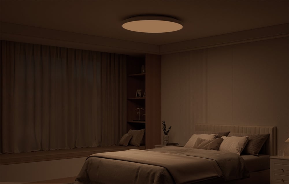 Przyciemnione wnętrze sypialni z łóżkiem i świecącą w ciepłym świetle lampą sufitową Yeelight Mercury C300.