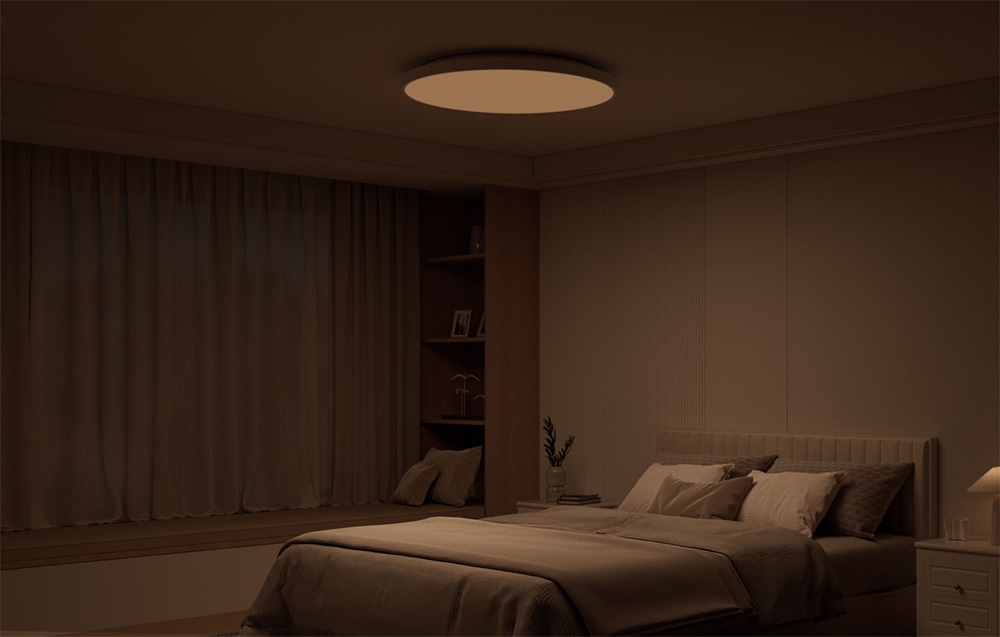 Przyciemnione wnętrze sypialni z łóżkiem i świecącą w ciepłym świetle lampą sufitową Yeelight Mercury C300.