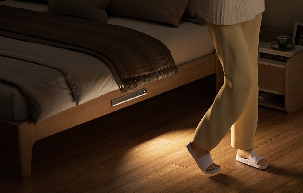 Lampka do szafy Yeelight zamontowana pod łóżkiem oświetlająca podłogę obok stojącej osoby.