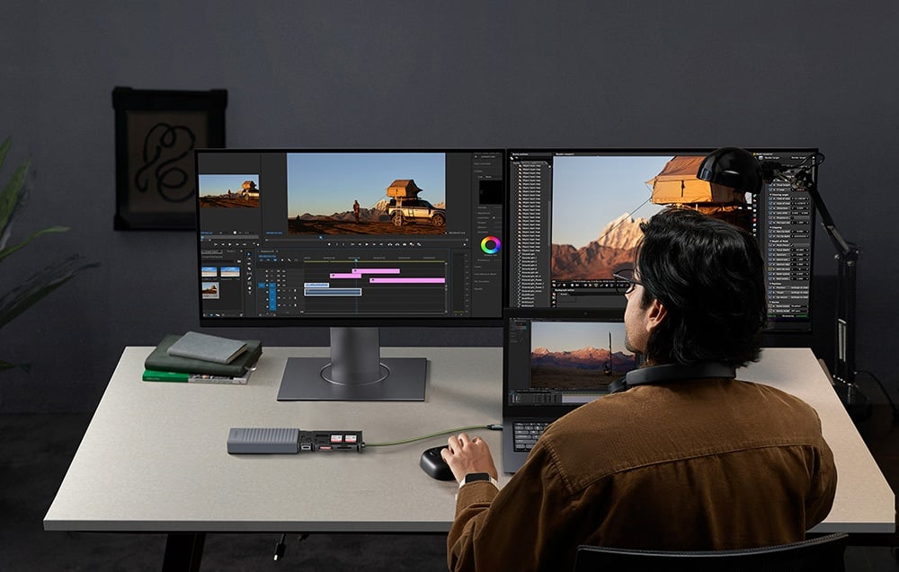 Mężczyzna pracuje przy biurku z trzema ekranami, edytując materiał wideo i korzystając z podłączonego hubu