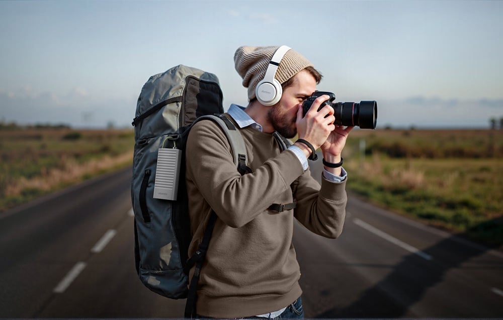 Mężczyzna z plecakiem i słuchawkami robi zdjęcie lustrzanką na poboczu drogi, z przyczepionym do plecaka hubem