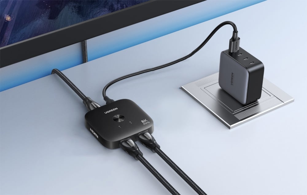 Przełącznik Ugreen AW700 leżący na biurku, podłączony do ładowarki sieciowej i monitora.