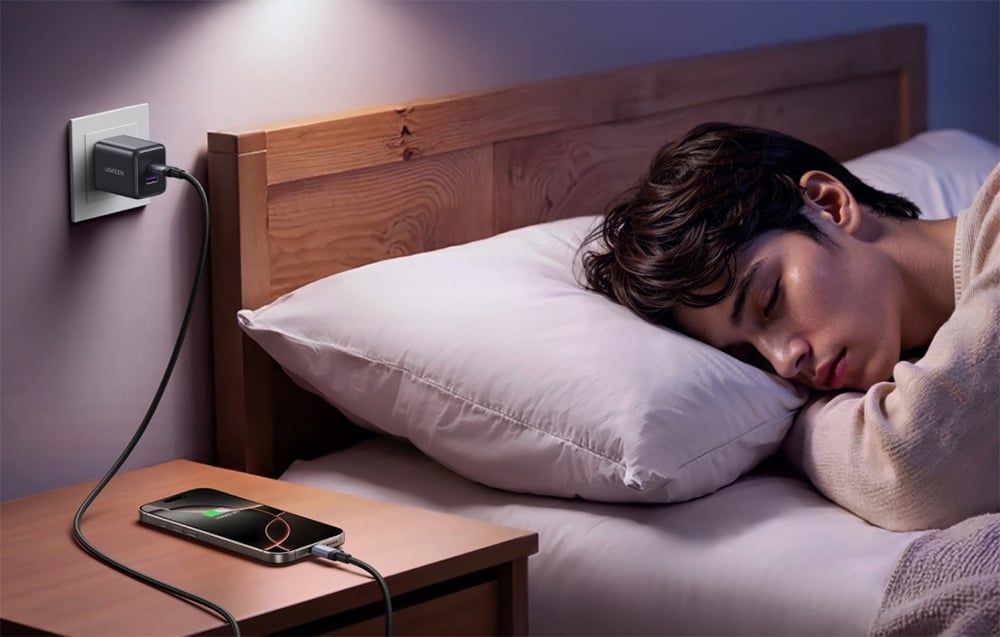 Mężczyzna śpiący w łóżku, obok na drewnianej półce stoi smartfon zasilany przez ładowarkę i kabel Ugreen L532.
